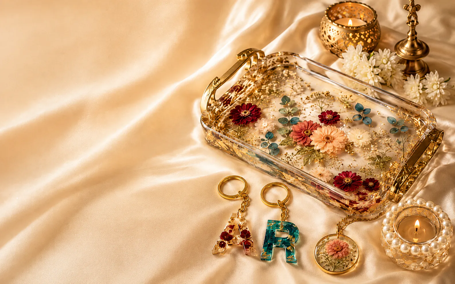 Floral resin tray, pearl diya holder, alphabet keychains and pendant arranged on champagne silk